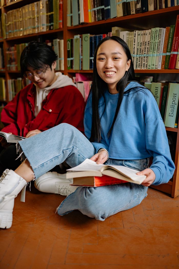 Two students sitting on the library floor reading books and smiling, capturing a moment of learning and friendship.