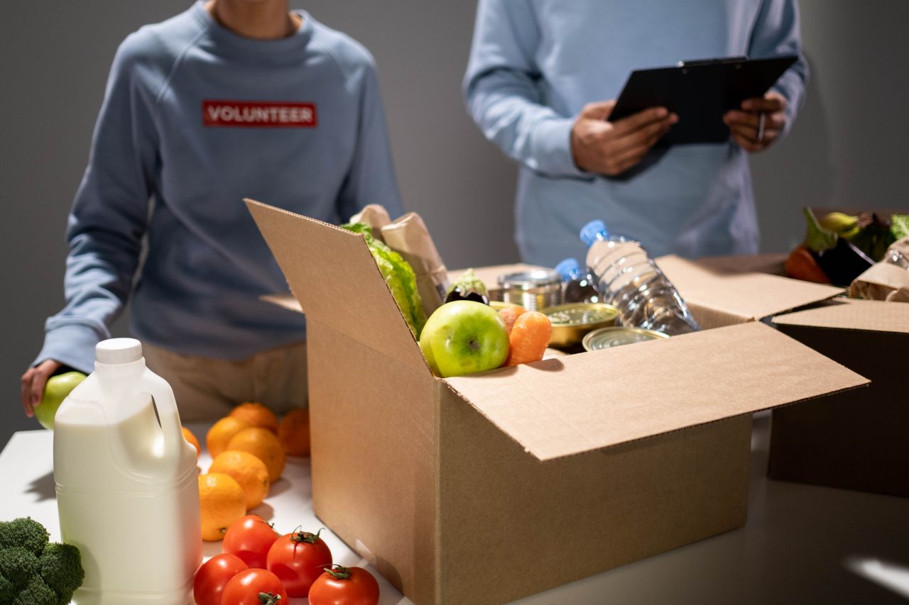 People packing a cardboard box with essentials like fruits, vegetables, and bottled water for charity.